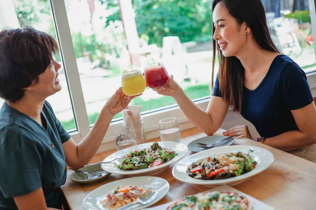 Side view of Asian female friends clinking glasses of fresh drinks while sitting at table with delicious food in restaurant